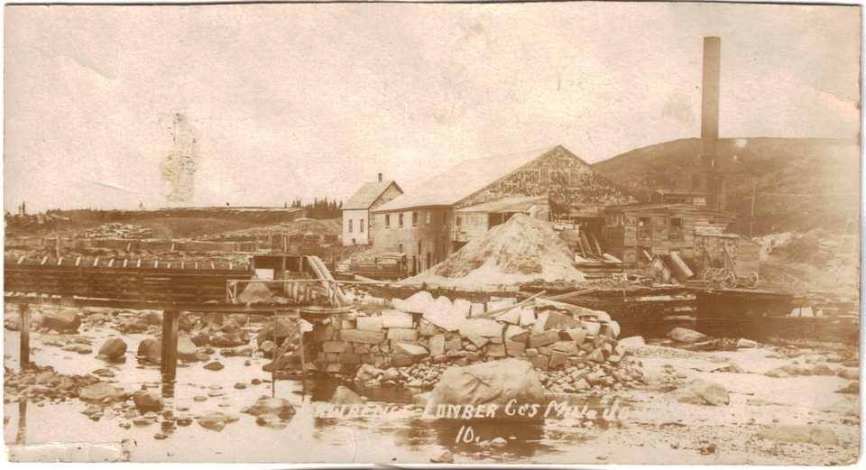 Sepia photograph of a riverside mill with a tall chimney, a sawdust pile and a stone wall, seen across a shallow, rocky river.