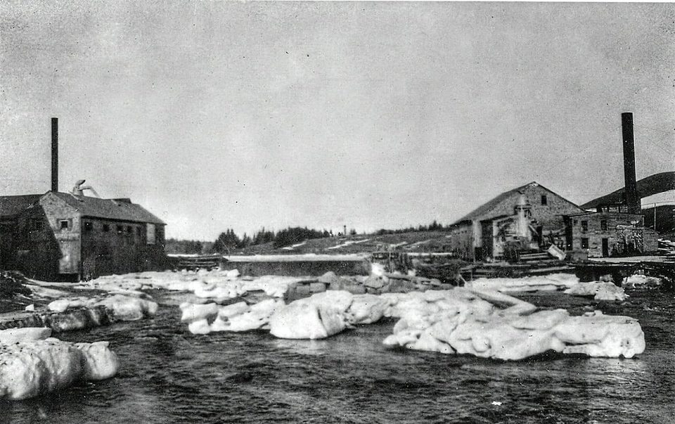 Black-and-white photograph of a river crowded with large blocks of ice, with a mill building and tall chimney on each bank.